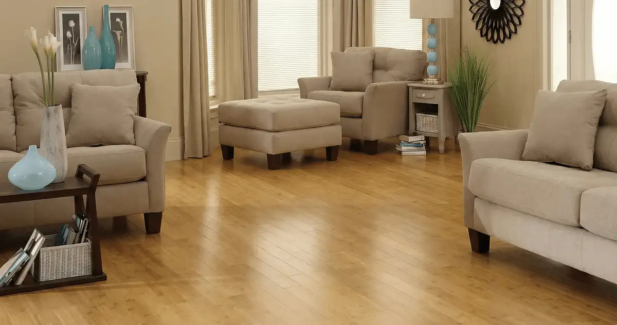 A living room with professionally installed bamboo flooring, beige sofas, a matching ottoman, a side table with a blue lamp, vases, framed pictures, and large windows with blinds and curtains in Savannah, GA